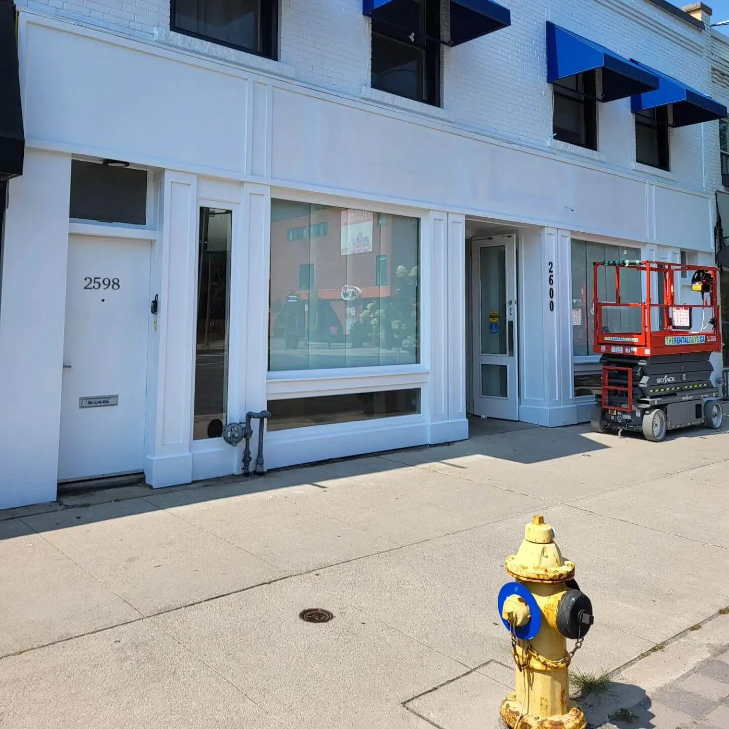 Commercial storefront with freshly painted white exterior, blue awnings, and a red scissor lift parked outside on a sunny day.