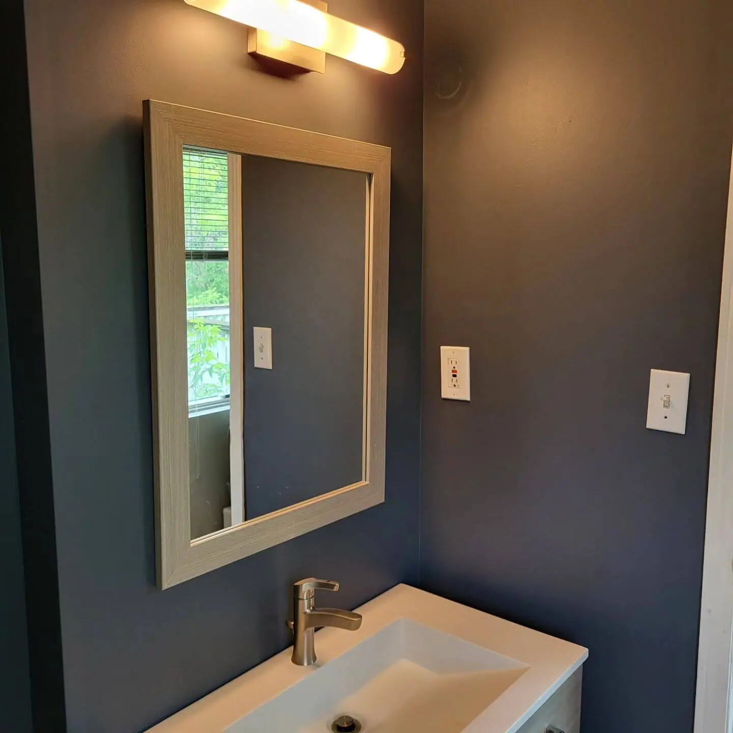 Modern bathroom vanity with a rectangular mirror, a single-handle faucet, and a wall-mounted light fixture above, complemented by dark gray walls. Modern bathroom vanity with a rectangular mirror, a single-handle faucet, and a wall-mounted light fixture above, complemented by dark gray walls.