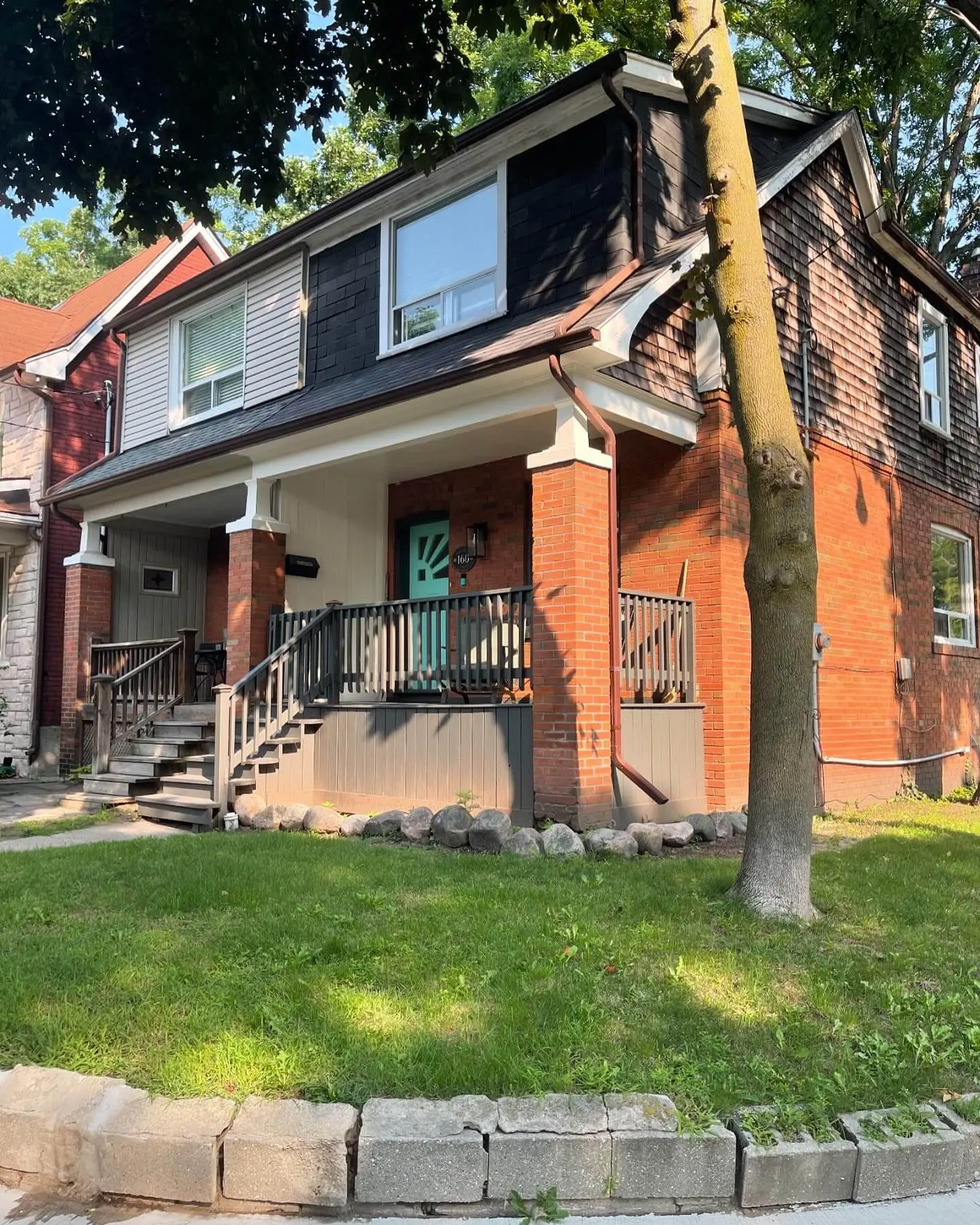 Front view of a two-story brick house with a porch, a turquoise front door, wooden railing, and a small front yard with a tree and lawn. Front view of a two-story brick house with a porch, a turquoise front door, wooden railing, and a small front yard with a tree and lawn.