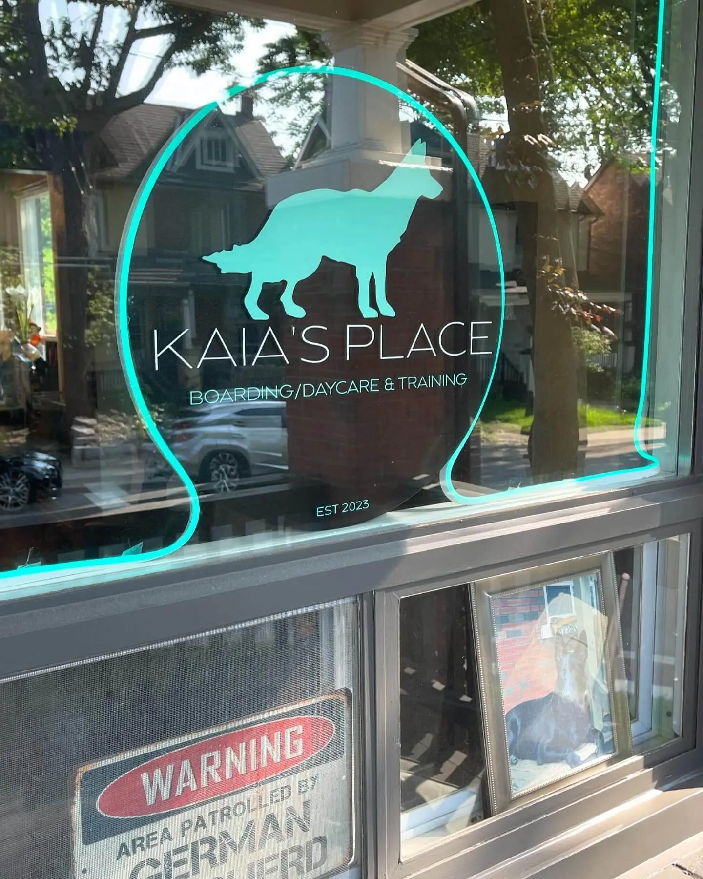 Glass window displaying the logo for 'Kaia's Place' with text 'Boarding/Daycare & Training' and a dog silhouette, framed by reflections of the neighborhood.