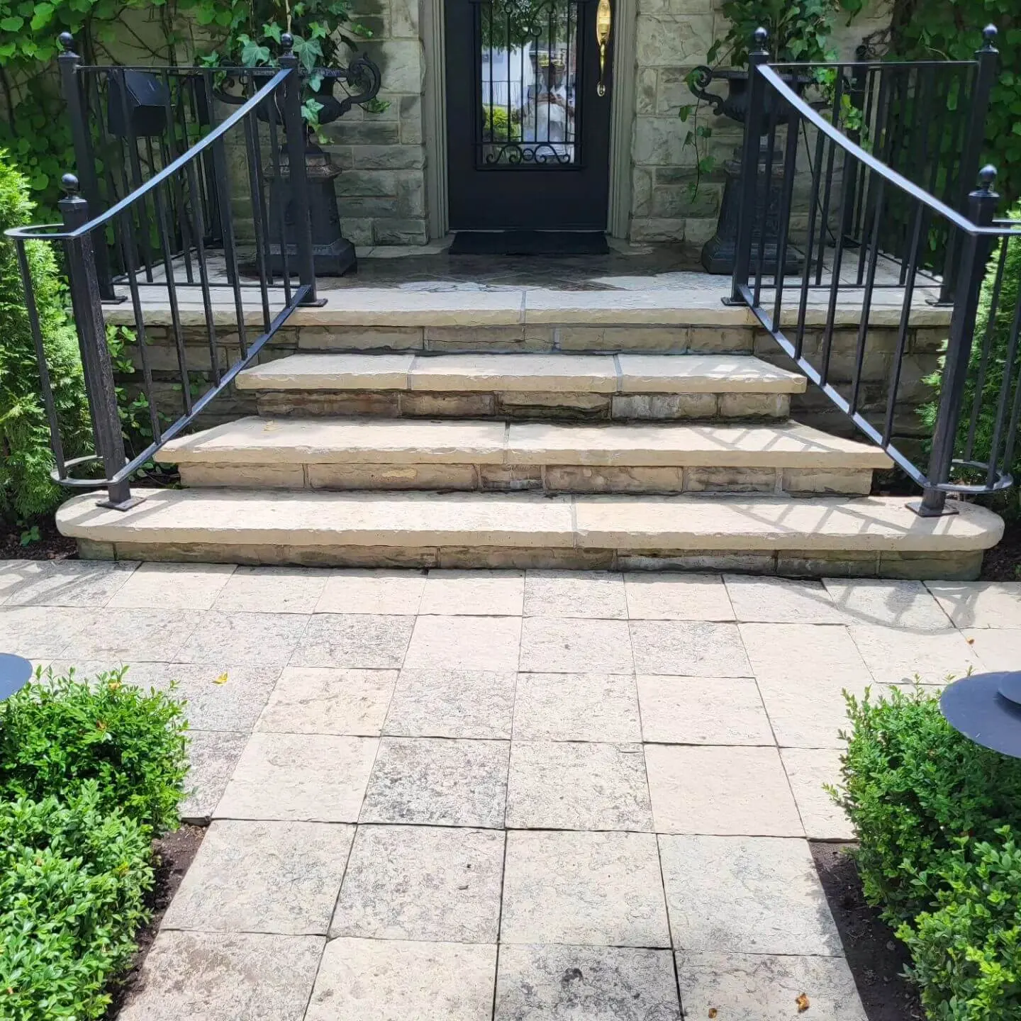 Elegant front steps with black iron railings leading to a stone doorway framed by climbing plants and greenery.