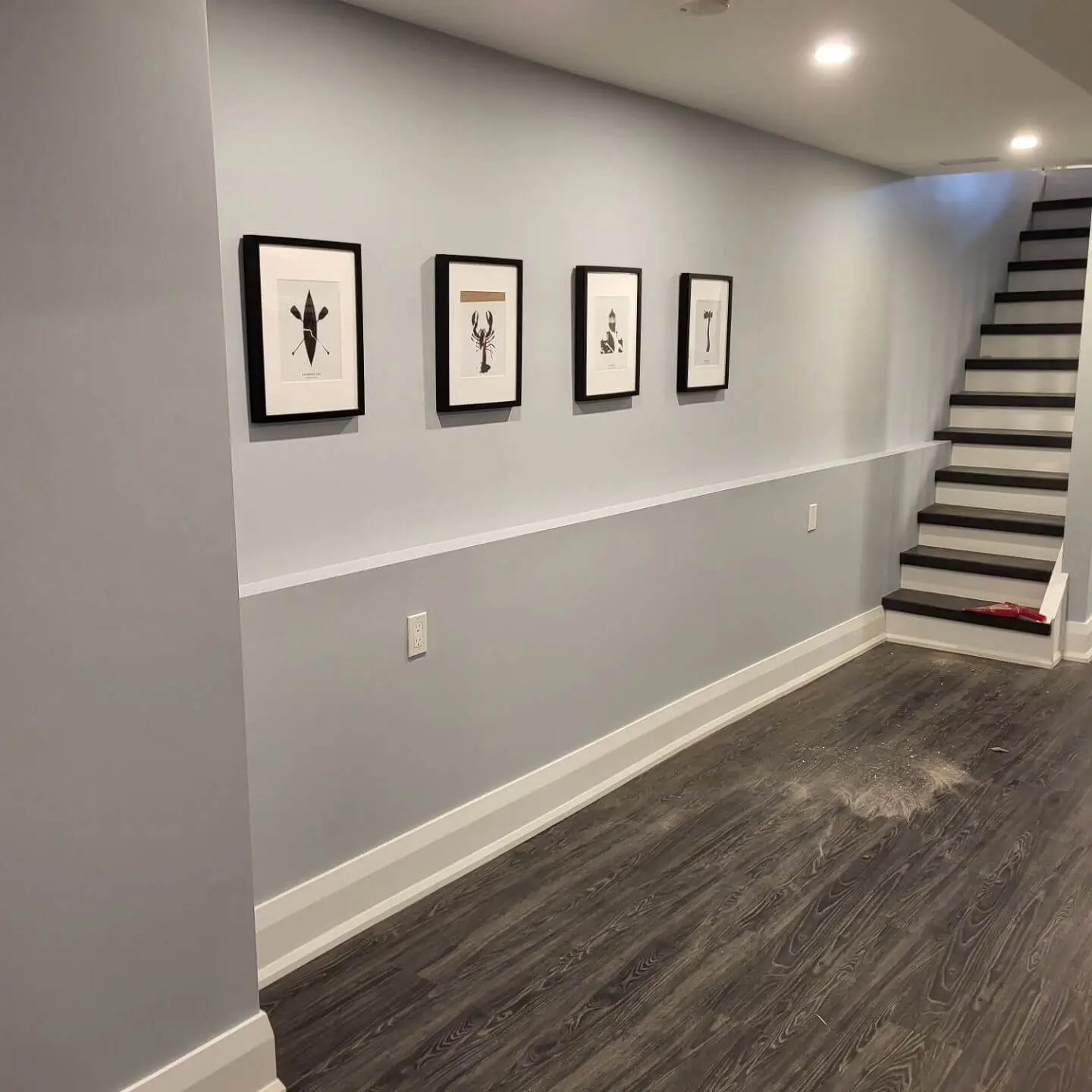 Modern finished basement with light gray walls, framed artwork, dark wood flooring, and a staircase leading upstairs.
