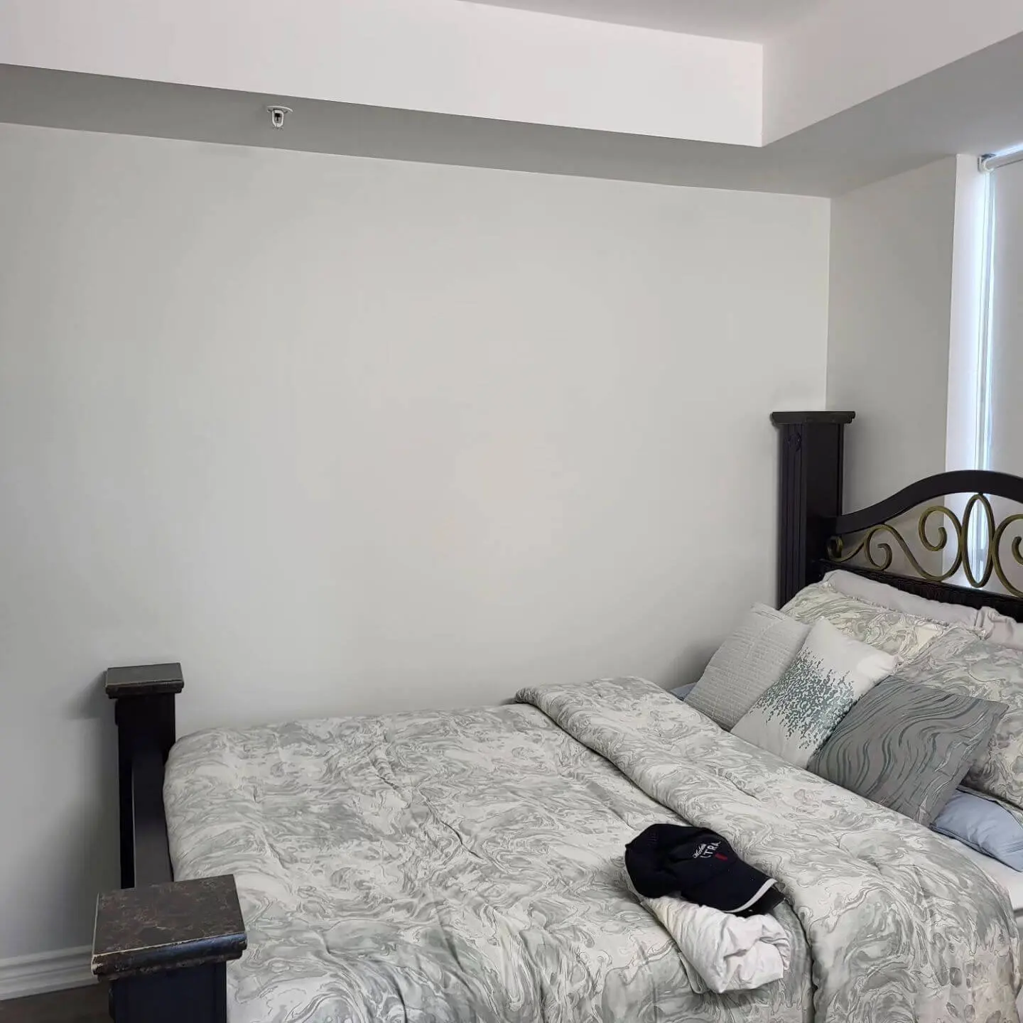 Bedroom with a black metal-framed bed, white bedding with light gray patterns, and a plain white wall in the background.