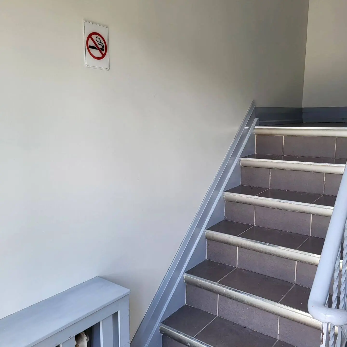 Indoor staircase with gray tiled steps, a light gray railing, and a 'No Smoking' sign on the white wall.