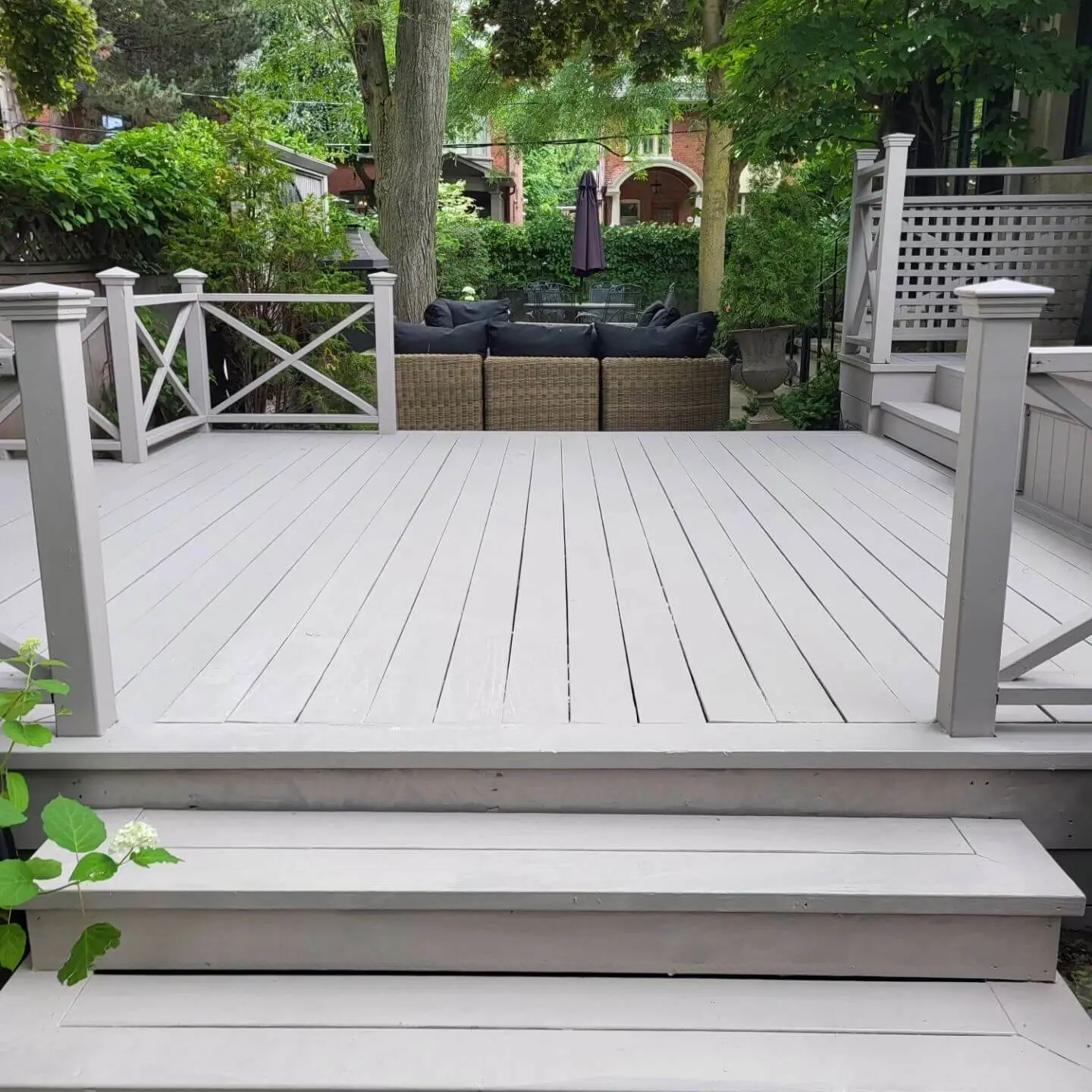 Freshly painted gray wooden deck with stairs, surrounded by lush greenery, leading to an outdoor seating area with wicker furniture.